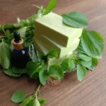 Organic Kuppaimeni Soap bars and a blue glass dropper bottle on a bed of fresh green leaves with "DM Traders Organic store" logo.