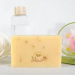 A Lemongrass Soap Bar With Dm Traders Organic Store Logo, Placed In Front Of Toiletries And Decorative Items.