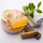 A Display Of Sandalwood Soap Skincare Products Including A Soap Bar, A Bowl Of Powder, A Small Bottle, Fresh Green Leaves, Wooden Pieces, And A White Towel, Branded By Dm Traders Organic Store.